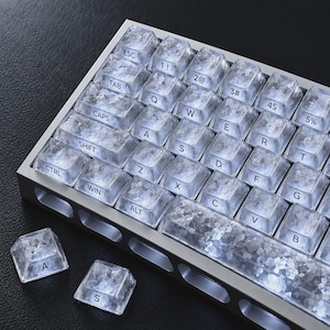 Puede incluir: Un teclado mecánico con marco plateado y teclas translúcidas con un diseño interior cristalino y texturizado. Cada tecla muestra letras negras, incluyendo caracteres QWERTY estándar, ESC, TAB, CAPS, SHIFT, CTRL, WIN y ALT.