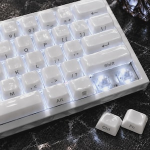 May include: A white mechanical keyboard with backlit keys featuring standard alphanumeric characters, symbols, and a Shift key. Two loose white keycaps labeled Ctrl and Fn are positioned separately next to the keyboard frame.