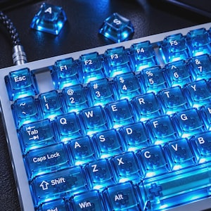 May include: A mechanical keyboard featuring translucent blue keycaps with white lettering and bright internal backlighting. The keys include standard QWERTY layout characters, function keys, and modifier keys like Shift, Tab, and Caps Lock, with two loose keycaps labeled A and S visible above the main board.