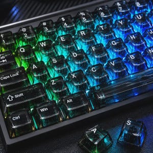 Può includere: Una tastiera meccanica con tasti traslucidi di tonalità scura e lettere bianche. La tastiera è illuminata da una vivace retroilluminazione a gradiente che sfuma dal verde a sinistra al blu a destra. Due tasti sfusi sono posizionati accanto.