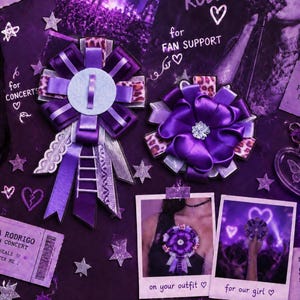 May include: Two purple ribbon rosettes featuring layered satin, leopard print accents, and a central silver embellishment. One rosette includes hanging ribbon tails with a small ladder charm. The set is displayed alongside concert-themed paper ephemera.