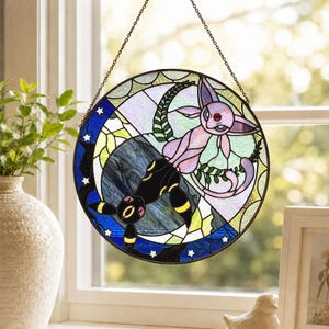 May include: A circular stained glass suncatcher featuring a split design with a light pink creature on a pale background and a black creature with yellow accents on a dark blue background, both surrounded by white stars and a metal chain.