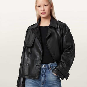 May include: A black leather jacket featuring a double-breasted front with four visible buttons, a wide collar, shoulder epaulets, and adjustable buckled straps at the cuffs. The jacket is worn over a plain black top and blue denim jeans.