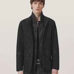 May include: A black jacket featuring a structured collar, a front button closure, and two flap pockets at the waist. The jacket is worn over a dark shirt and dark denim jeans, with a black belt featuring a silver buckle visible at the waistline.