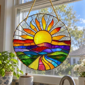 Può includere: Un acchiappasole circolare in vetro colorato raffigurante un paesaggio vivace con un sole giallo brillante, raggi arancioni e colline ondulate in tonalità di rosso, viola, blu e verde. L'oggetto è incorniciato in metallo nero e appeso a un cordino dorato.
