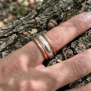 Peut inclure: Un ensemble de bagues empilables composé de trois anneaux lisses et arrondis. L'anneau central est en métal argenté poli, encadré par deux anneaux plus fins de couleur cuivre, porté sur un doigt devant une écorce texturée.