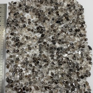 May include: A large collection of small, translucent, and dark-speckled quartz crystals arranged in a rectangular shape next to a metal ruler that shows the collection spans approximately 10 centimeters in height.