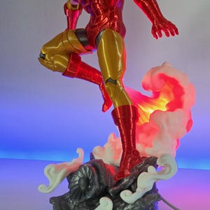 Könnte beinhalten: Eine metallisch rote und goldene Iron Man Actionfigur in dynamischer Flugpose, montiert auf einem dunkelgrauen felsigen Sockel mit weißen skulptierten Raucheffekten. Der Sockel verfügt über eine Lichtquelle, die den Rauch warm beleuchtet.