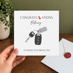 Peut inclure: Une carte de vœux carrée blanche avec le texte CONGRATULATIONS, le prénom Bethany et ON PASSING YOUR DRIVING TEST! La carte présente un dessin de clé de voiture et d'une télécommande avec une étiquette DRIVE SAFE! Un L rouge remplace le U.