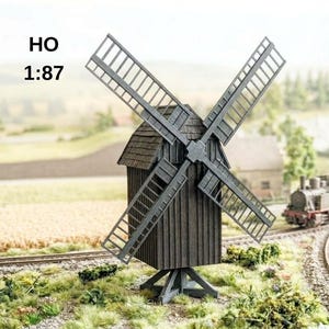 Pode incluir: Um moinho de vento em miniatura cinza-escuro com quatro pás em treliça e um telhado de telhas, sobre uma base de vigas cruzadas. O modelo inclui o texto HO 1:87, indicando a sua escala para maquetes de comboios e dioramas.