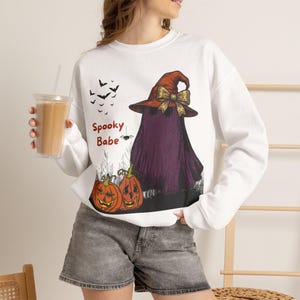 Peut inclure: Un sweat-shirt blanc à manches longues orné d'un motif représentant un fantôme violet portant un chapeau de sorcière marron avec un nœud doré, deux citrouilles orange, des chauves-souris noires et le texte rouge Spooky Babe, avec un short en jean gris.