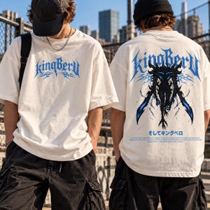 May include: An off-white short-sleeve t-shirt featuring a blue KingBerU logo on the chest. The back displays a large, detailed black and blue graphic of a creature with swords, the text KingBerU, and Japanese characters.