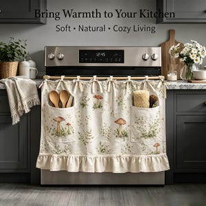 May include: A beige fabric kitchen stove curtain featuring a mushroom and greenery print, two front pockets, and a ruffled bottom hem. The curtain is tied to the stove handle with fabric bows and contains the text Bring Warmth to Your Kitchen.