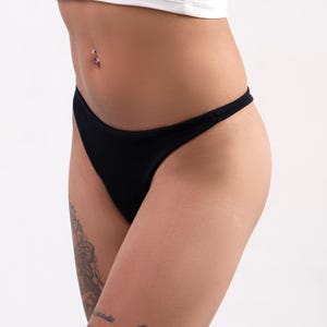 Könnte beinhalten: Ein schwarzer Bikini-Slip mit dünnen Seitenträgern, getragen zu einem weißen gerippten bauchfreien Oberteil. Der Slip hat einen klassischen Schnitt und bietet eine schlichte und klare Ästhetik für den täglichen Bedarf an Unterwäsche.