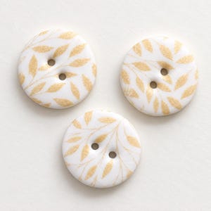 May include: Three round white buttons featuring a gold leaf pattern. Each button has two centered holes for attachment. The surface of the buttons is smooth and circular, displaying a repeating botanical print in a metallic gold color.