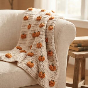 Puede incluir: Una manta de ganchillo de color crema con un patrón repetitivo de pequeñas calabazas naranjas en relieve con tallos verdes. La manta tiene un diseño de punto acanalado texturizado y está colocada sobre el brazo de un sofá tapizado claro.