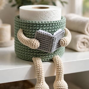 Puede incluir: Un soporte para papel higiénico de ganchillo en color verde salvia con un diseño de personaje con brazos y piernas beige. La figura sostiene un pequeño libro de ganchillo gris en sus manos. El soporte es cilíndrico para un rollo de papel blanco.