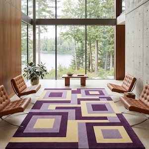 Puede incluir: Una alfombra rectangular con un patrón geométrico de cuadrados y rectángulos entrelazados en tonos morado oscuro, lavanda claro y amarillo pálido. La alfombra tiene una textura de pelo corto y uniforme y cubre una gran parte del suelo.