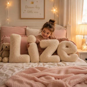 May include: Four cream-coloured, textured, plush letter-shaped pillows spelling the name LIZE. Each letter is crafted from a soft, fuzzy fabric and is positioned on a bed. The pillows are arranged in a row, showcasing a rounded, chunky design.