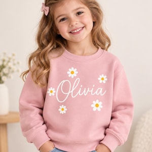 May include: A light pink long-sleeve sweatshirt featuring a crew neckline and ribbed cuffs. The front of the garment displays the name Olivia in white cursive script, surrounded by several white daisy graphics with yellow centers.