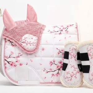 Könnte beinhalten: Eine weiße Pferdesatteldecke mit rosa Kirschblütenmuster, eine passende rosa gehäkelte Fliegenhaube und zwei weiße Gamaschen mit demselben Blumenmuster, schwarzen Riemen und einem weichen, cremefarbenen Kunstfellfutter.