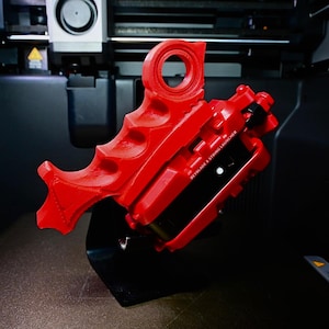 Peut inclure: Un lanceur de ficelle Beyblade X rouge vif imprimé en 3D, doté d'une poignée ergonomique avec des rainures pour les doigts et d'une boucle supérieure circulaire. Le texte BEYBLADE X STRING LAUNCHER est gravé en blanc sur le côté du dispositif.