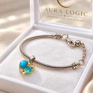 May include: A silver-tone snake chain bracelet featuring a heart-shaped charm filled with turquoise-coloured stones and gold-coloured flakes. The bracelet is displayed in a white box with the text AURA LOGIC MEMORIES THAT LAST FOREVER.