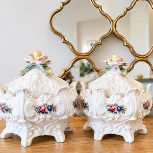 May include: Two white ceramic lidded bowls featuring intricate embossed scrollwork and a scalloped base. Each lid is topped with a sculpted pink and yellow flower. The sides are decorated with small floral clusters in red, blue, and green.