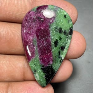 Peut inclure: Une pierre précieuse polie en forme de poire présentant un mélange vibrant de rubis rouge magenta profond et de zoïsite vert lime vif, agrémentée d'inclusions de hornblende noire dispersées sur toute la surface, avec une finition lisse et brillante.