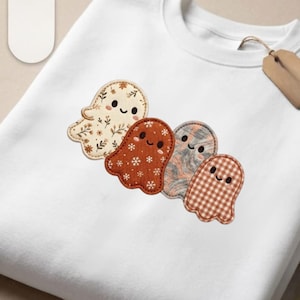 Könnte beinhalten: Ein weißes Sweatshirt mit Rundhalsausschnitt und einer Stickerei von vier kleinen, in einer Reihe angeordneten Geistern. Jeder Geist hat ein eigenes Muster: einer mit Blumenmotiven, einer mit weißen Schneeflocken auf Rost, einer mit grauer Marmorierung und einer mit braunem Vichy-Karo.