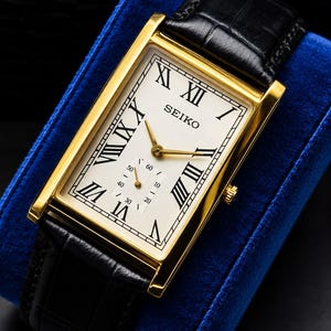 May include: A rectangular Seiko wristwatch with a gold-tone case, a white dial featuring black Roman numerals, and gold-tone hands. The watch includes a sub-dial for seconds and a black leather strap with a textured, embossed pattern on the surface.