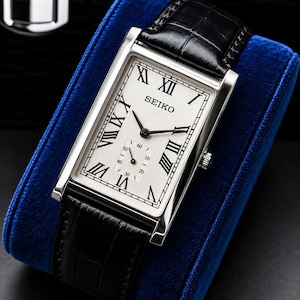 May include: A rectangular Seiko wristwatch with a silver-toned dial, black Roman numeral hour markers, and a small sub-dial for seconds. The watch features a polished silver-colored metal case and a black textured leather strap with a croc-embossed finish.