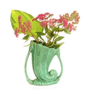 May include: A green ceramic vase shaped like a seashell with two handles. The vase is filled with pink flowers and green leaves.