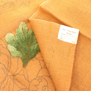 May include: A close-up of a partially completed rug hooking project featuring a green leaf design. The rug hooking is on a tan canvas with a label that reads "Pea's Craft House Designs".