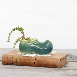 May include: A small green ceramic shoe planter with a succulent plant growing out of it. The planter is sitting on top of an old book with the title "Sweet Clover" visible.