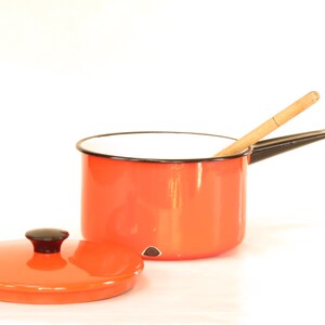 May include: Orange enamel saucepan with a black handle and a wooden spoon inside. The saucepan has a matching orange lid.