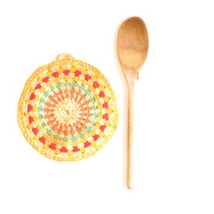 May include: A round, crocheted pot holder with a colorful, concentric circle pattern in shades of red, yellow, green, and orange. A wooden spoon is placed next to the pot holder.