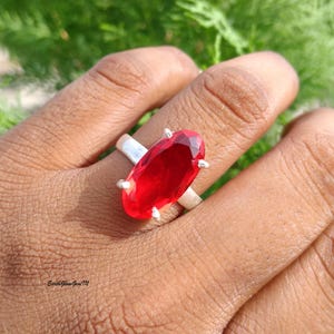 May include: A silver ring featuring a large, oval-cut, vibrant red gemstone held in place by four prongs. The band is smooth and metallic, showcasing a polished finish. The text EarthGlowGemTM is visible in the lower left corner of the image.