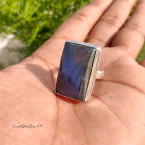 May include: A rectangular labradorite gemstone ring set in a silver-toned metal band. The stone features a deep blue iridescent flash across its surface. The ring is displayed on a palm, and the text EarthGlowGemIN is visible in the corner.