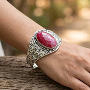 Peut inclure: Un bracelet manchette argenté orné d'une pierre précieuse rouge foncé ovale et facettée en son centre. La bande métallique présente des motifs de volutes en relief complexes et une bordure perlée entourant le sertissage central.