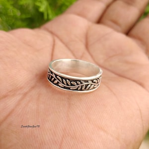 May include: A silver band ring featuring a continuous embossed pattern of small, stylised leaves encircling the outer surface. The metal has a polished finish on the edges with darkened, recessed areas highlighting the detailed leaf design.