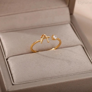 Puede incluir: Un anillo abierto de tono dorado que presenta una letra A mayúscula en un extremo y una gema redonda transparente en el otro. El anillo tiene una banda fina y pulida, presentado en un joyero de color neutro con el texto Elyrune visible.