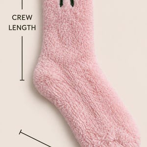 Puede incluir: Un calcet&iacute;n de color rosa claro y textura afelpada de longitud media con un peque&ntilde;o lazo bordado en verde oscuro en la parte superior. La imagen incluye etiquetas de texto que indican CREW LENGTH y ONE SIZE FITS MOST.