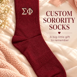 Puede incluir: Un calcetín de caña media acanalado de color granate con las letras griegas Sigma Phi bordadas en hilo blanco. El texto CUSTOM SORORITY SOCKS y A big-little gift to remember está impreso en granate junto al calcetín sobre tela blanca.
