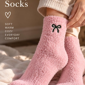 Puede incluir: Un par de calcetines rosas difusos con un peque&ntilde;o detalle de lazo bordado en verde oscuro en el tobillo. El texto en la imagen dice: Fuzzy Socks, s&iacute;mbolo de coraz&oacute;n, SOFT, WARM, COZY, EVERYDAY, COMFORT. Los calcetines tienen un acabado texturizado.