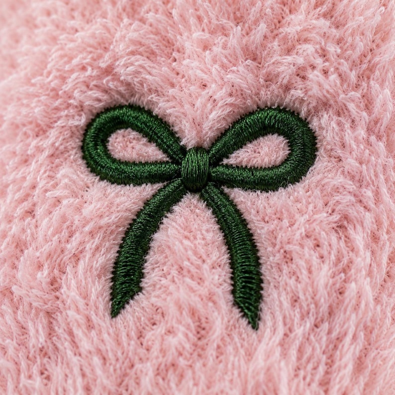 Puede incluir: Una tela suave y difusa de color rosa claro que presenta un lazo bordado de color verde oscuro en el centro. La textura del material es afelpada y peluda, lo que proporciona un alto contraste visual con las puntadas densas y lisas del lazo.
