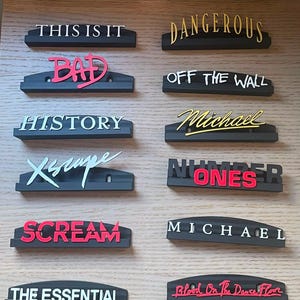 May include: A collection of twelve black rectangular display stands, each featuring raised lettering in various colors and fonts. The text includes Thriller, Invincible, This Is It, Dangerous, Bad, Off The Wall, History, Michael, Xscape, Number Ones, Scream, The Essential, and Blood On The Dance Floor.