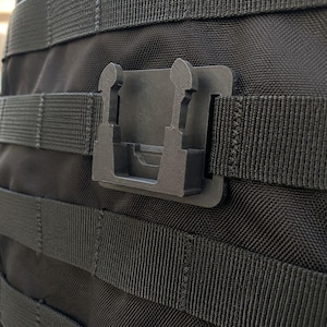 Pode incluir: Um clipe de montagem de plástico preto fosco preso a uma mochila tática de nylon preta com tiras horizontais. O clipe apresenta uma base retangular com duas abas de travamento verticais projetadas para prender equipamentos ao sistema MOLLE.