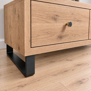 Peut inclure: Un meuble télé en bois clair avec un grain apparent, doté d'un tiroir rectangulaire et d'un bouton circulaire unique. Le bouton présente un centre sombre et un contour doré. L'ensemble repose sur un pied en métal noir mat en forme de U.