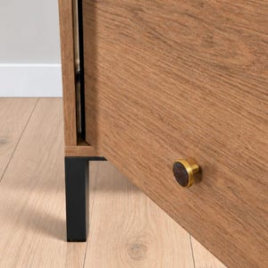 May include: A wooden cabinet door with a visible grain pattern, featuring a circular knob with a dark centre and a metallic gold rim. The cabinet is supported by a rectangular black metal leg, showing a clean edge and a smooth finish.